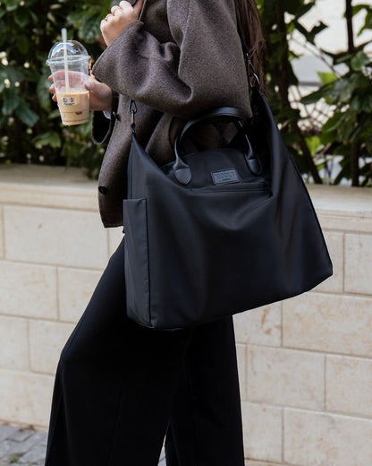 Person holding a black shoulder bag and a coffee cup outdoors.