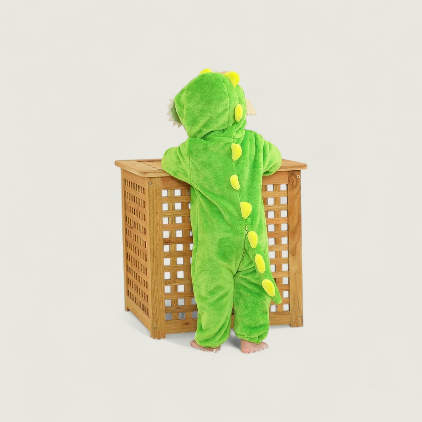 Child wearing a green dinosaur onesie standing next to a wooden basket on a light gray background