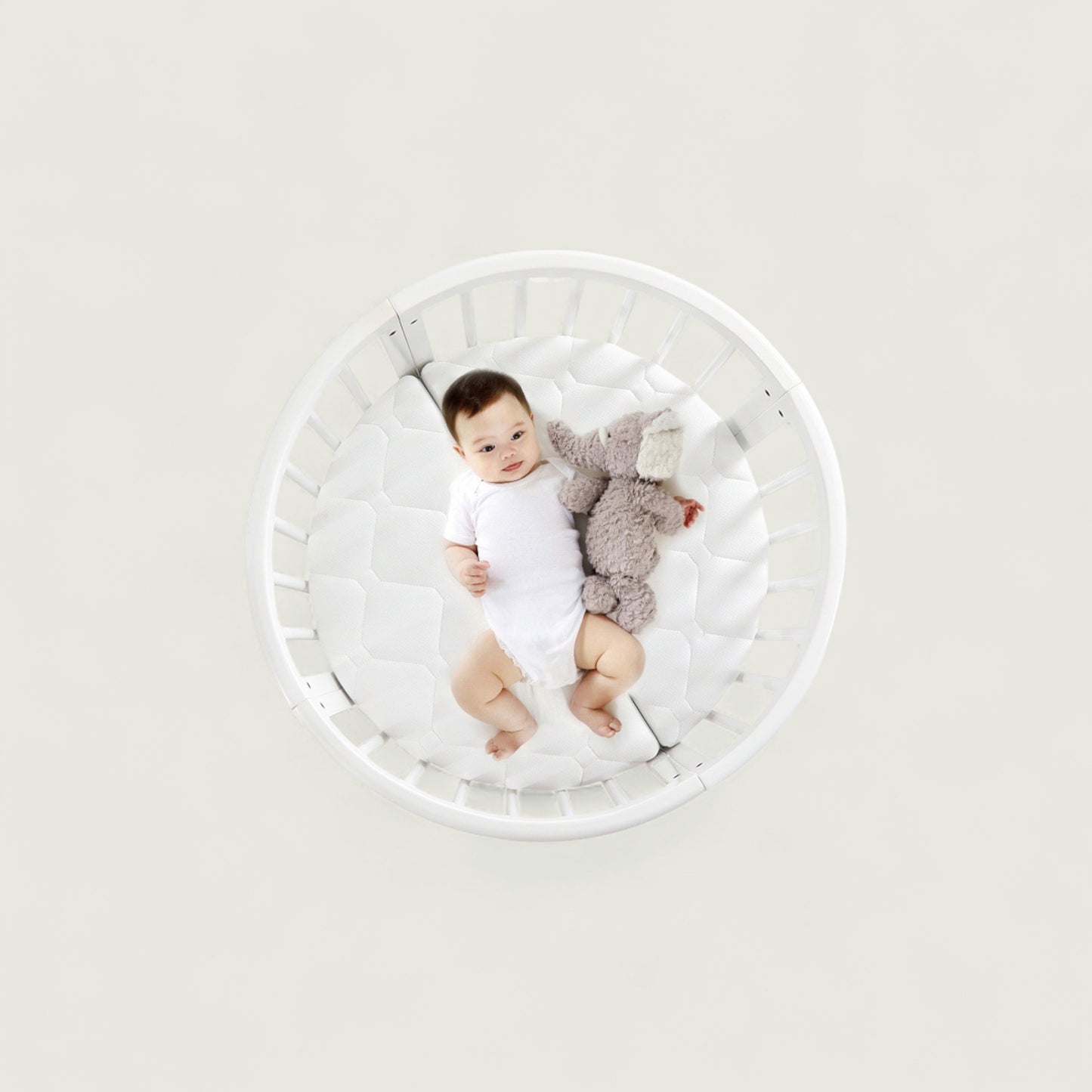 Baby lying in a white crib with a gray toy on a light gray background