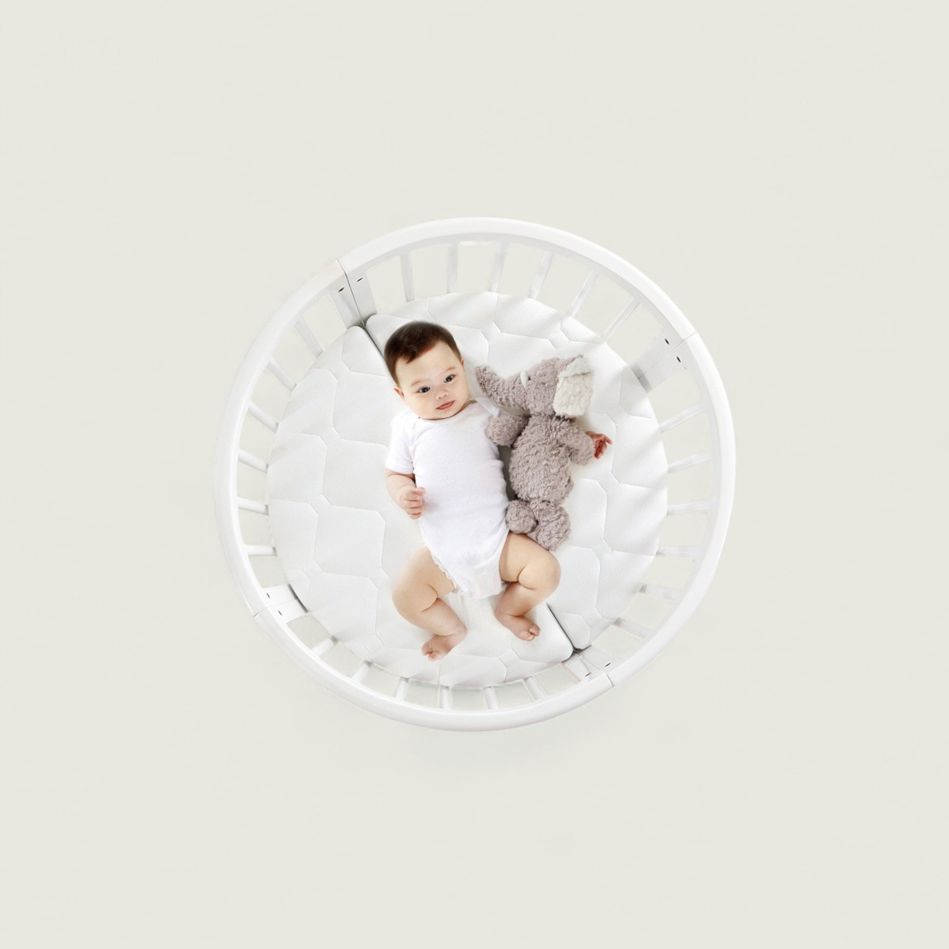 Baby lying in a white crib with a gray toy on a light gray background