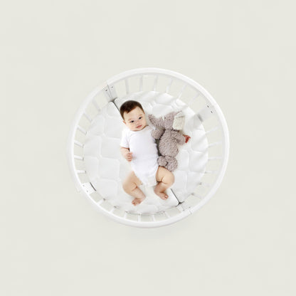 Baby lying in a white crib with a gray toy on a light gray background