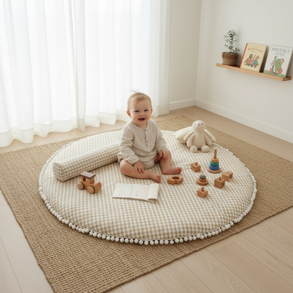 Baby sitting on a round cushion with toys in a room with white curtains and a shelf.