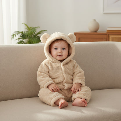 Baby in a beige bear onesie sitting on a couch in a bright room.