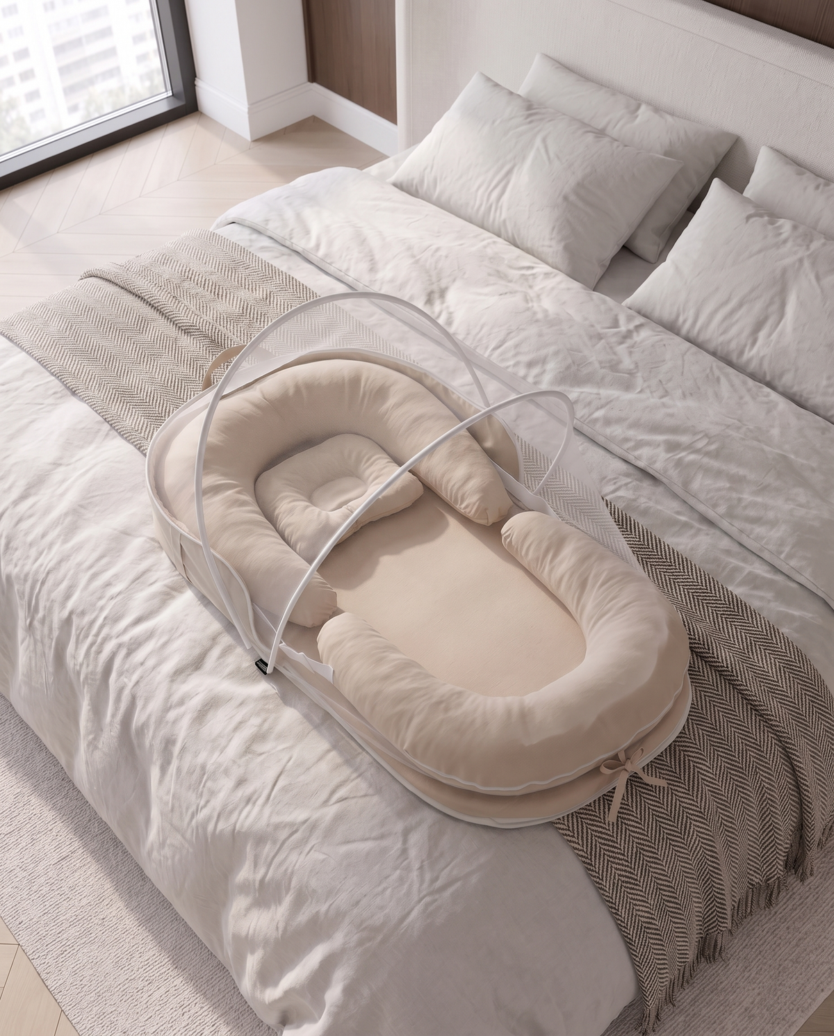 A beige baby bassinet with a white mesh mosquito net cover is set up on a neatly made bed with white linens and a textured beige throw blanket.