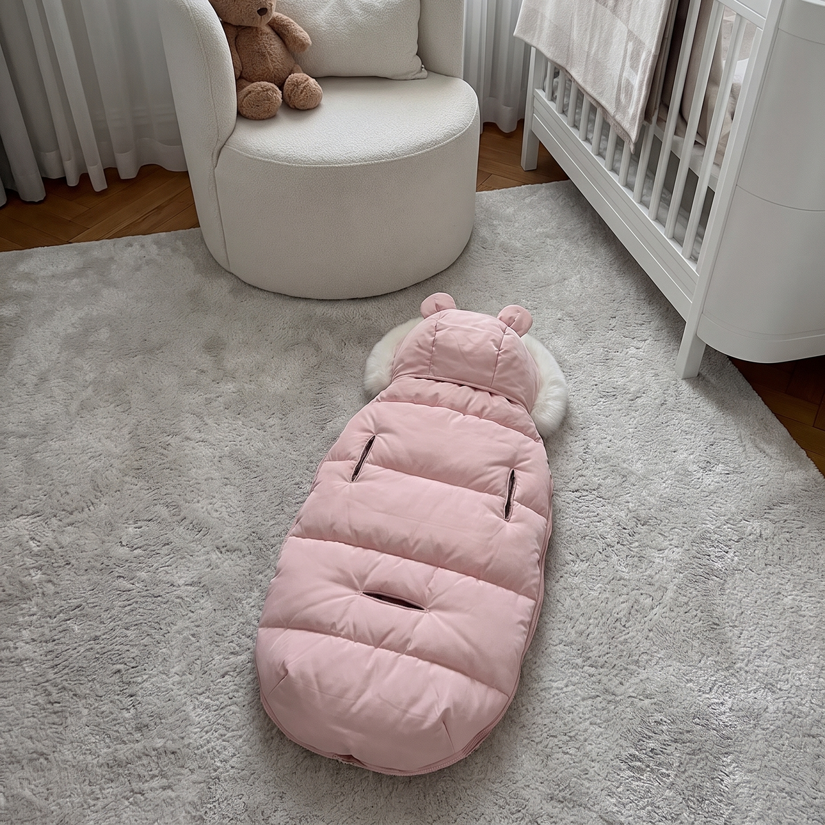 A pink quilted baby ground cover with a hood and ear details lies on a light gray rug in a nursery setting, with a white armchair, teddy bear, and white crib visible in the background.