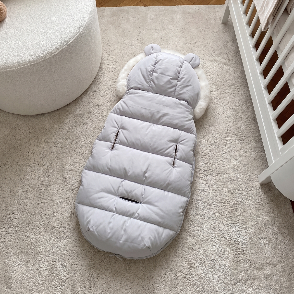 A light gray puffer baby blanket with a hood and ear details, laid out on a beige rug in a nursery setting next to a white crib and chair.