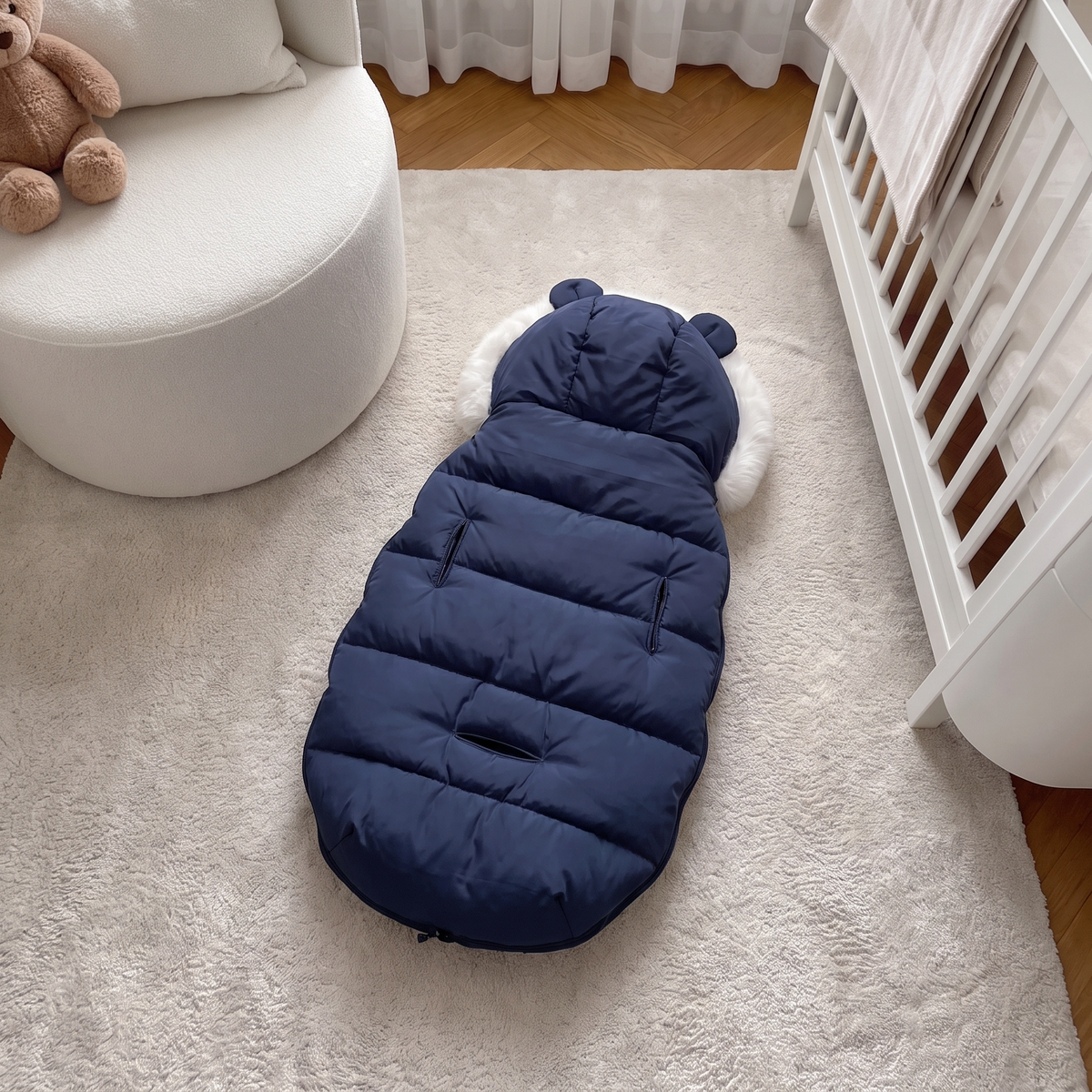 A navy blue baby sleeping bag or footmuff with a white faux-fur trim, laid out on a white rug in a modern nursery setting next to a white crib and chair.