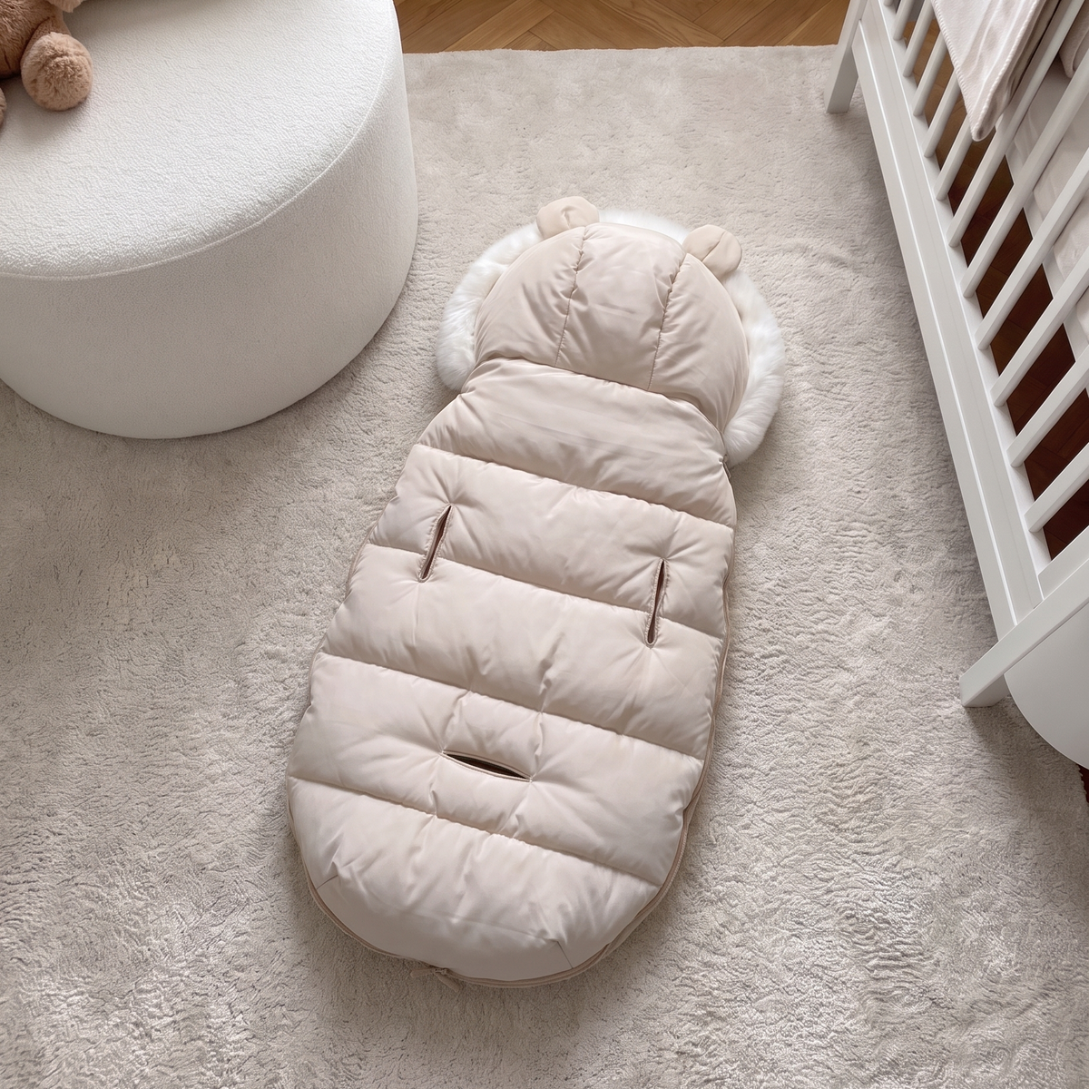A cream-colored puffer baby ground mat with ear details on the hood, laid out on a fluffy cream rug in a nursery setting next to a white crib and chair.