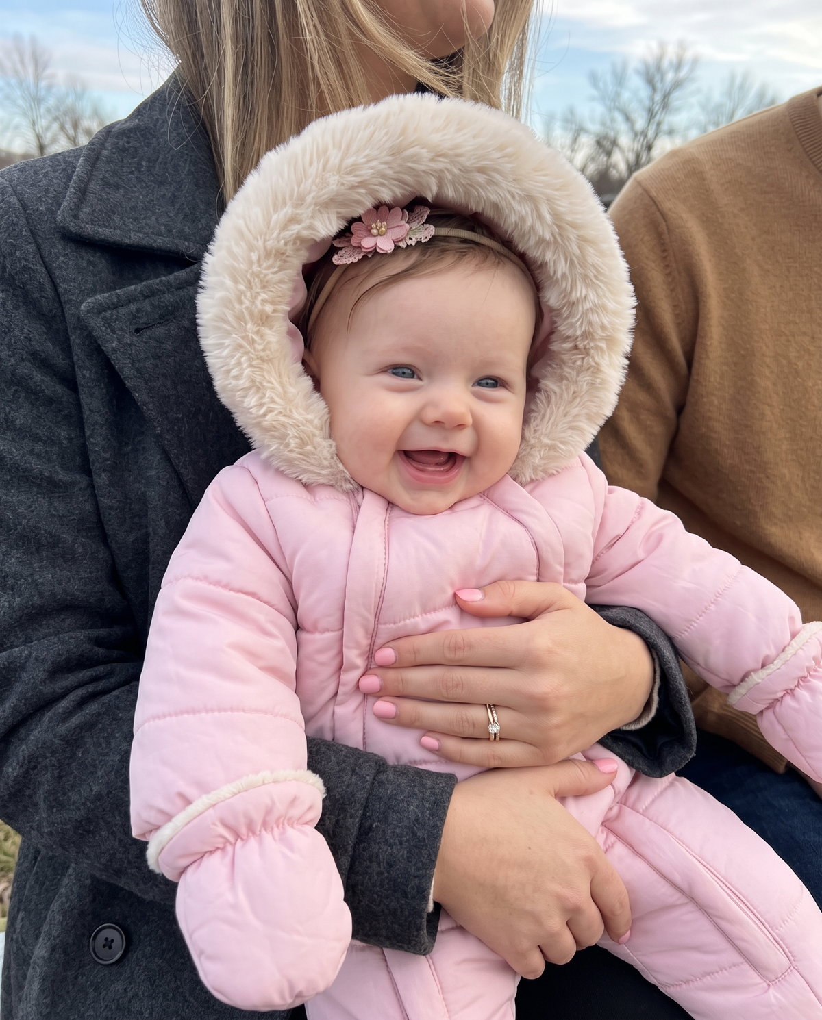 A baby wearing a pink winter snowsuit with a fluffy beige hood and a pink flower headband, being held by an adult outdoors.