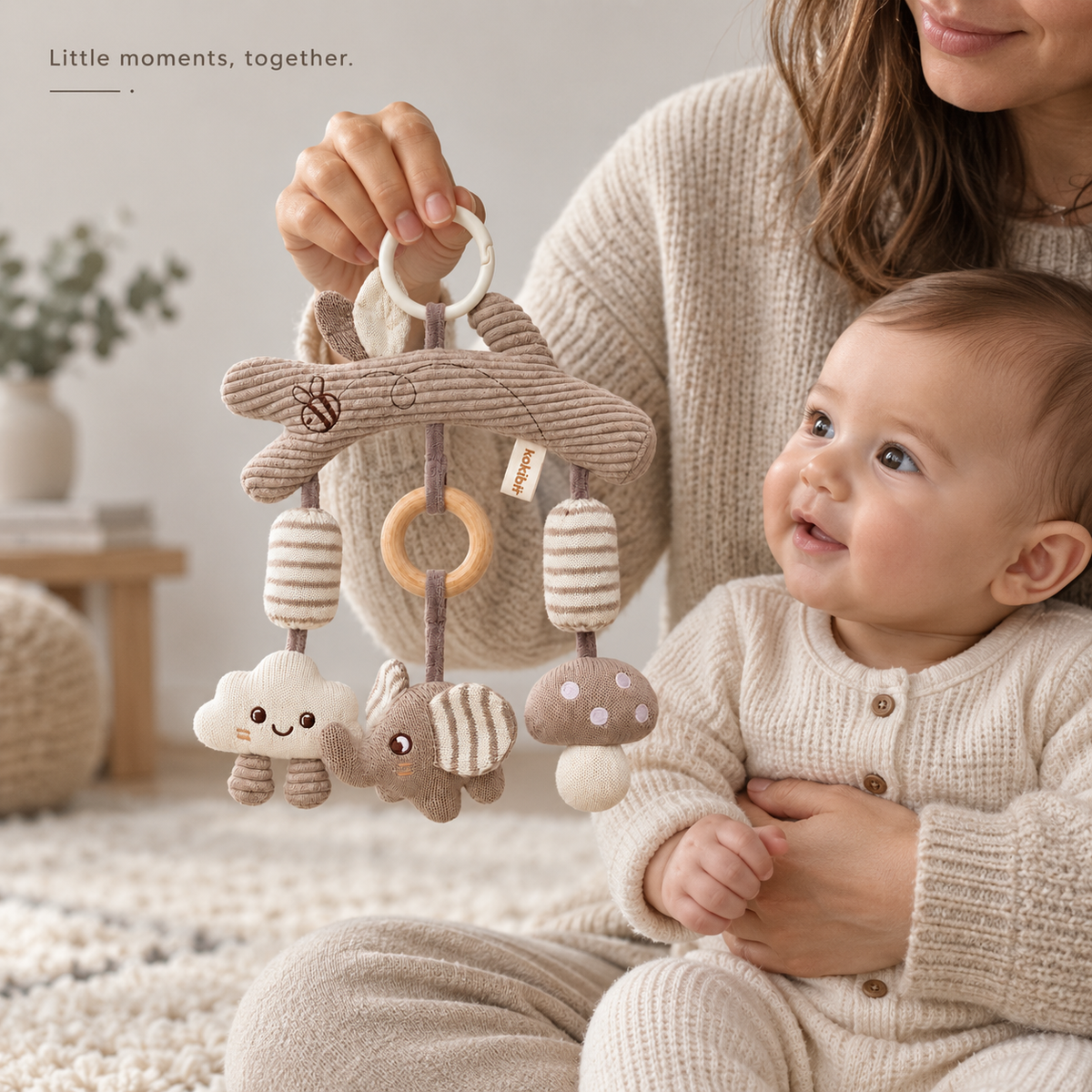 Adult holds the cloud, elephant and mushroom hanging toy while an awake baby watches. Little moments, together.